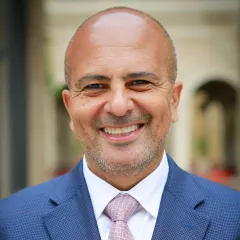 Headshot of a smiling man with medium skin tone and close-shaved head wearing white shirt, purple tie and blue suit against a blurred indoor background