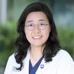 Headshot of a smiling woman with light skin tone and shoulder-length dark hair wearing round, black wire-rimmed glasses and a white lab coat against a blurred outdoor background