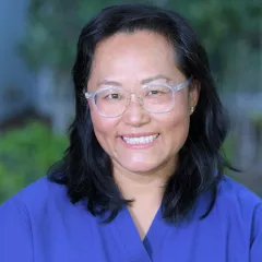 Headshot of a smiling woman with light skin tone and dark shoulder-length hair wearing clear framed glasses and a blue top against a blurred outdoor background