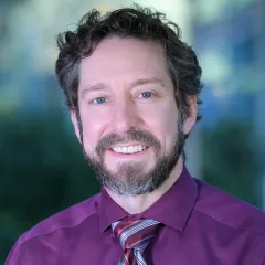 Headshot of a smiling, bearded man with light skin tone and dark hair wearing a purple shirt and red and grey striped tie against a blurred outdoor background