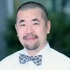 Headshot of a smiling man with light skin tone, closely shaved dark hair and a goatee wearing a white, checkered dress shirt and plaid bow tie against a blurred outdoor background