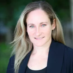 Headshot of a smiling woman with light skin tone and shoulder-length blonde hair wearing a dark suit jacket against a blurred outdoor background