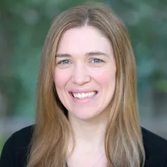 Headshot of a smiling woman with light skin tone and light brown hair wearing a black top against a blurred outdoor background