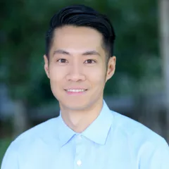 Headshot of smiling young man with medium skin tone and dark hair wearing button-down blue dress shirt against a blurred outdoor background