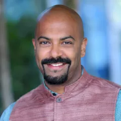 Headshot of a smiling man with dark skin tone and a goatee wearing a purple vest against a blurred outdoor background