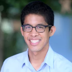 Headshot of a smiling man with medium skin tone and dark hair wearing tortoise shell glasses and an open-collar light blue dress shirt against a blurred outdoor background