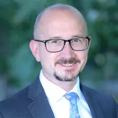 Headshot of a smiling man with light skin tone, shaved head and a goatee, wearing dark rimmed glasses, a white dress shirt and tie under a dark suit jacket against a blurred outdoor background