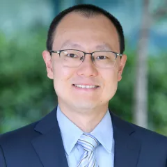 Headshot of a smiling man with medium skin tone and short dark hair wearing glasses, a blue dress shirt and striped tie under a dark suit jacket against a blurred outdoor background