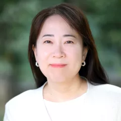 Headshot of a woman with a slight smile, light skin tone and dark hair, wearing a white top under a white suit jacket against a blurred outdoor background