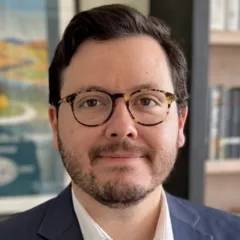 Headshot of a smiling man with short brown hair, a light beard and tortoise-shell glasses wearing an open-collar, white dress shirt under a dark blue suit jacket against a blurred indoor background