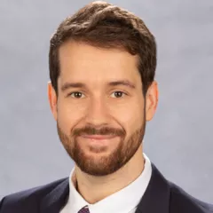 Headshot of a smiling man with medium skin tone, short brown hair and a closely cropped beard wearing a white dress shirt under a dark suit jacket against a neutral indoor background