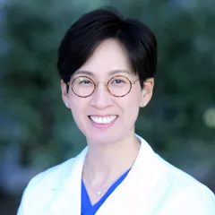 Headshot of a smiling woman with light skin tone and short dark hair wearing round tortoise shell glasses and a blue blouse under a white lab coat against a blurred outdoor background