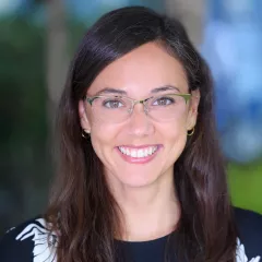 Headshot of a smiling woman with light skin tone and long dark hair wearing eyeglasses, small gold hoop earrings and dark blouse with white floral print against a blurred outdoor background