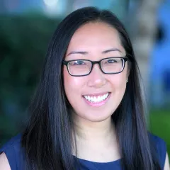 Headshot of a smiling woman with light skin tone and straight dark hair wearing black-rimmed eyeglasses and a dark blue top against a blurred outdoor background