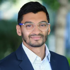 Headshot of a smiling man with medium skin tone and short dark hair wearing clear rimmed eyeglasses and a light patterned dress shirt under a dark suit jacket against a blurred outdoor background