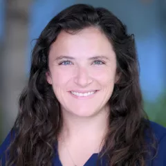 Headshot of a smiling woman with light skin tone and long wavy brown hair wearing a blue top against a blurred outdoor background