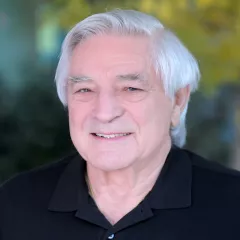 Headshot of a smiling man with light skin tone and short grey hair wearing a black polo shirt against a blurred outdoor background