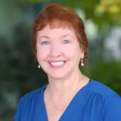 Headshot of a smiling woman with light skin tone and short, auburn hair wearing pearl earrings, a silver necklace and a blue, v-neck dress against a blurred outdoor background