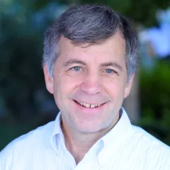 Headshot of a smiling man with light skin tone and grey hair wearing a light colored, open collared dress shirt against a blurred green outdoor background