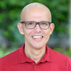 Headshot of a smiling man with medium skin tone and a shaved head wearing grey framed eyeglasses and a red polo shirt against a blurred green outdoor background