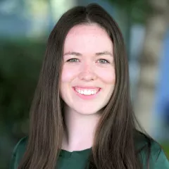 Headshot of a smiling woman with long brown hair and light skin tone wearing a green top against a blurred outdoor background