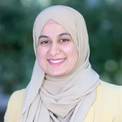 Headshot of a smiling woman with light skin tone wearing a beige headscarf and yellow suit jacket against a blurred outdoor background