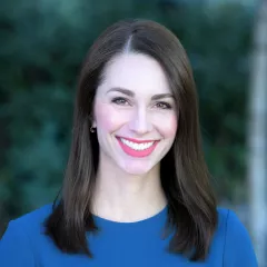 Headshot of a smiling woman with light skin tone and shoulder-length brown hair wearing a solid blue top against a blurred outdoor background