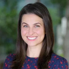 Headshot of smiling woman with light skin tone and long brown hair wearing blue top with red flower print against a blurred outdoor background