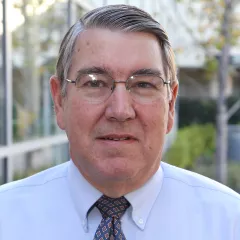An outdoor headshot of man with light skin and short grey hair, wearing thin-framed eyeglasses, a light blue dress shirt and dark blue patterned tie
