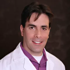 Headshot of a smiling man with light skin tone and dark hair wearing a purple shirt under a white lab coat against a neutral indoor background
