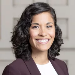 Headshot of a smiling woman with medium skin tone and shoulder-length, dark, wavy hair wearing a white blouse under a dark purple jacket against a neutral indoor background