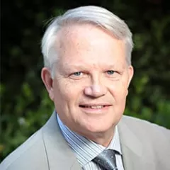 Headshot of a smiling man with light skin tone and grey hair wearing a striped dress shirt and silver tie under a grey suit jacket against a blurred green outdoor background