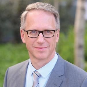 Headshot of Dr. David Freyer, a man with light skin tone and light hair wearing glasses and business attire with green bushes in the background