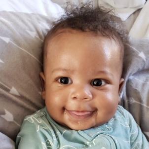 Smiling baby with dark skin tone and dark hair dressed in light green onesie resting on grey pillow patterned with white arrows