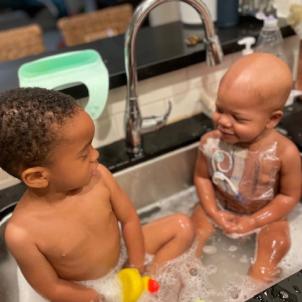 A toddler with dark skin tone and dark hair and an infant with dark skin tone share a bath together in a kitchen sink