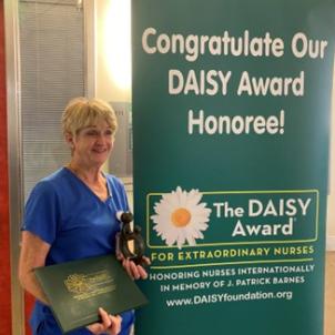 A female nurse with light skin tone and short blonde hair wearing blue hospital scrubs poses with their DAISY Award