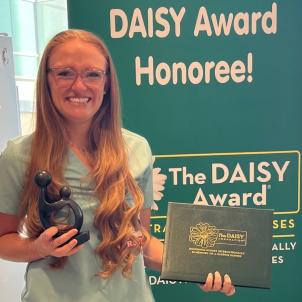Woman with light skin tone and long auburn hair wearing glasses and nurses' scrubs smiles while holding her DAISY Award
