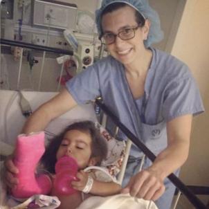 Smiling female surgeon with light skin tone wearing scrubs and glasses holds young girl's pink arm cast as she rests in hospital bed