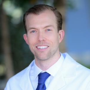 Headshot of smiling man with light skin tone and brown hair wearing a blue dress shirt and blue tie under a white lab coat against a blurred outdoor background