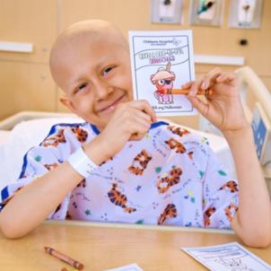 Young patient with medium skin tone and wearing a hospital gown with a tiger pattern smiles as they sit in hospital bed holding a CHLA Halloween card and a crayon