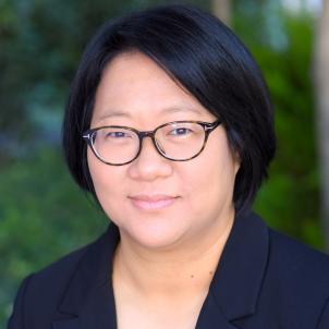 Headshot of a smiling woman with medium skin tone and short, dark hair wearing tortoise shell glasses and a dark suit jacket against a blurred outdoor background