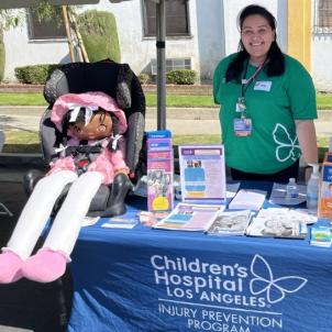 A medium skin toned woman smiles behind a table filled with safety information and equipment, including a life-sized doll in a child safety seat.