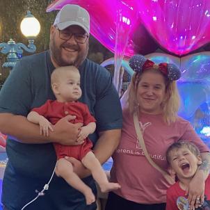 Father and mother with light skin tone pose for picture with toddler and young boy in front of colorful fountain at Disneyland