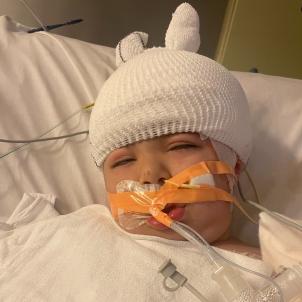 Young boy with medium skin tone with heavily bandaged head and breathing and feeding tubes lies against pillow in hospital bed