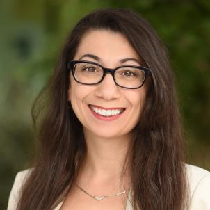 Headshot of a smiling woman with light skin tone and long brown hair wearing black framed eyeglasses, gold necklace and a cream colored suit jacket against a blurred outdoor background