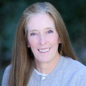 Headshot of a smiling woman with light skin tone and greying brown hair wearing a lavender cardigan against a blurred outdoor background