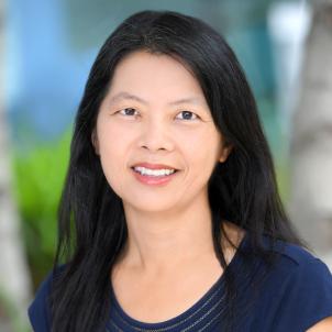 Headshot of a smiling woman with medium skin tone and shoulder-length dark hair wearing a blue top against a blurred outdoor background