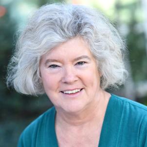 Headshot of a smiling woman with grey hair wearing a teal colored top against a blurred outdoor background