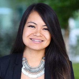 Headshot of a smiling young woman with medium skin tone and long dark hair wearing a large silver necklace and dark suit jacket against a blurred outdoor background