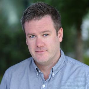 Headshot of a man with light skin tone and short brown hair wearing a open-collar blue dress shirt against a blurred outdoor background
